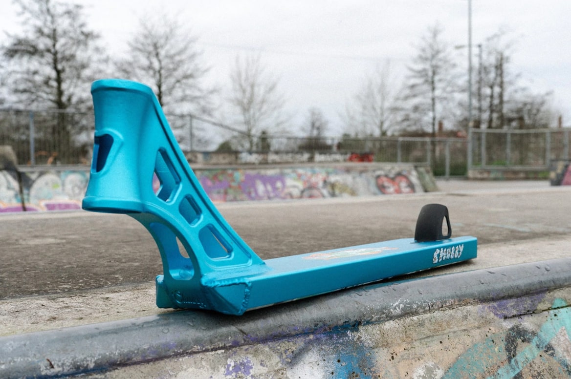 A top-notch Chubby Loco Marshmallow Boy Blue Stunt Scooter Deck, measuring 4.5" x 19.5", is positioned on a concrete surface in a skatepark adorned with graffiti. In the background, tall leafless trees contrast against the cloudy sky, providing an ideal atmosphere for executing exciting tricks and stunts.