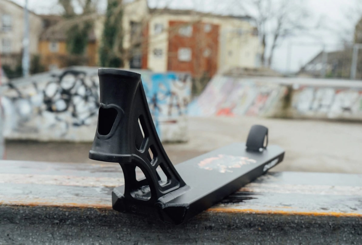A Chubby Loco Serpent Black Stunt Scooter Deck, measuring 4.5" x 19.5", with its lightweight and cutout designs, rests on a metal surface in an outdoor skate park. The blurred background showcases ramps adorned with graffiti and trees.