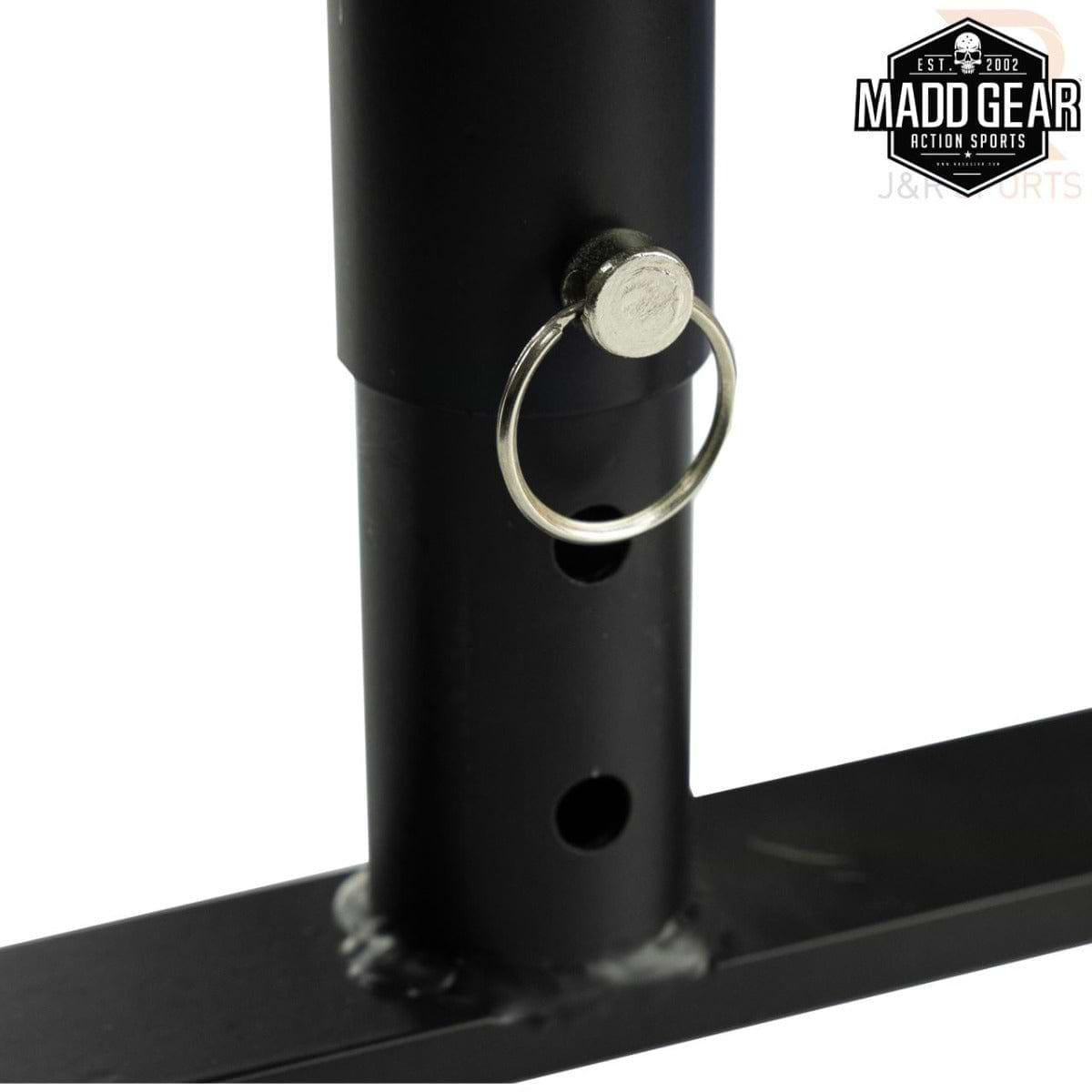 Close-up of the Madd Gear MGP Fifty5 Grind Rail - 140cm shows a locking pin inserted into one of its holes on the black metal rod, essential for perfecting grind tricks. This rail is part of a larger metal structure set against a white background, with the "Madd Gear" logo in the top right corner hinting at the adrenaline rush from skateboarding and more.
