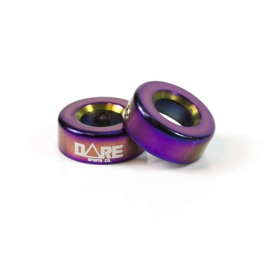 Two vibrant metal bushings with a lustrous, iridescent finish similar to the Dare CNC Aluminium Scooter Bar Ends - Neochrome showcase a gradient of purple, blue, and gold shades. One stands upright while the other rests on its side, prominently featuring the "DARE SPORTS CO." logo.