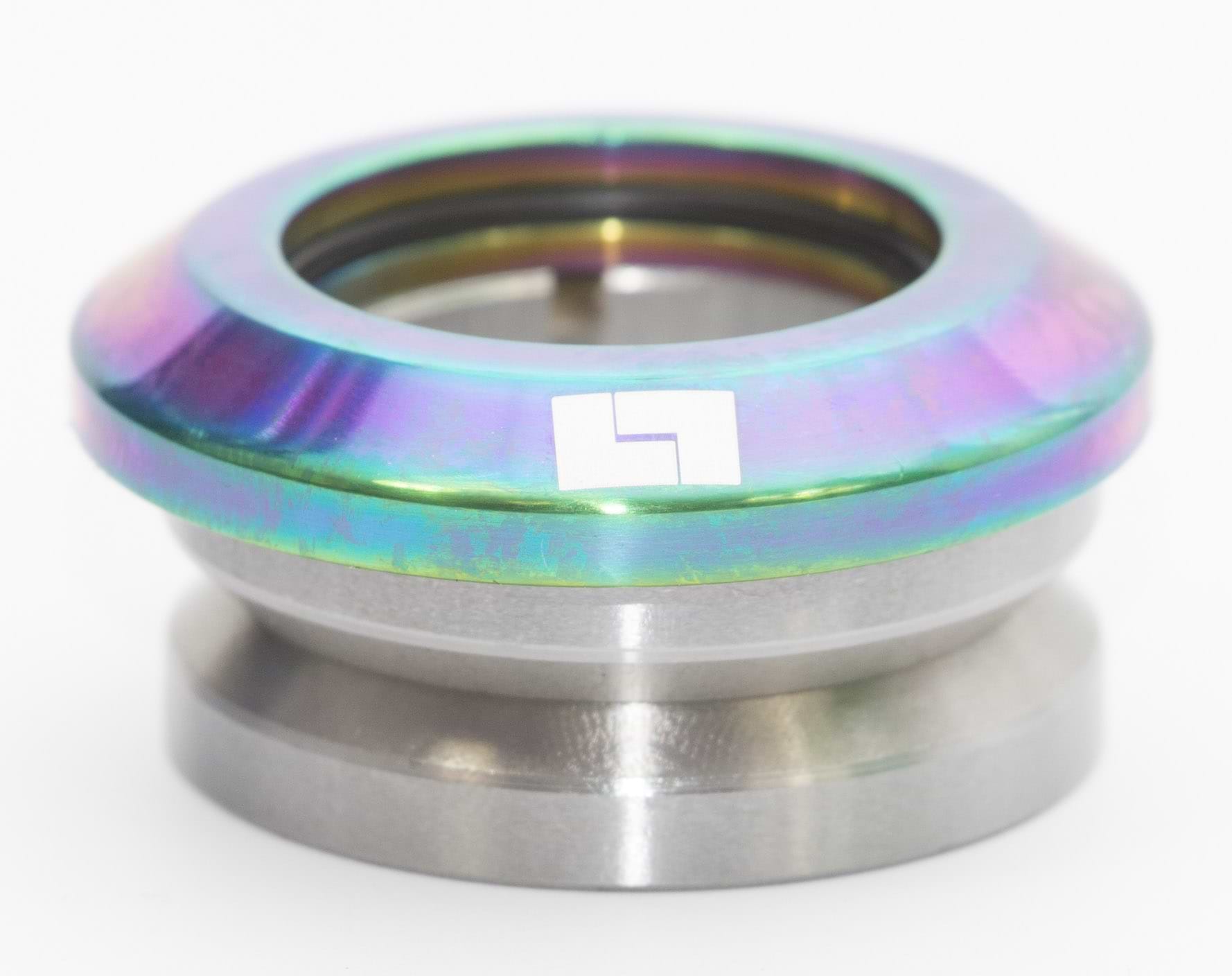 A component from the Logic Integrated Stunt Scooter Headset in Neochrome, showcasing a metallic, circular design with an iridescent finish similar to stylish scooter forks. The surface includes a small white square adorned with a black geometric pattern, all presented on a simple white background.