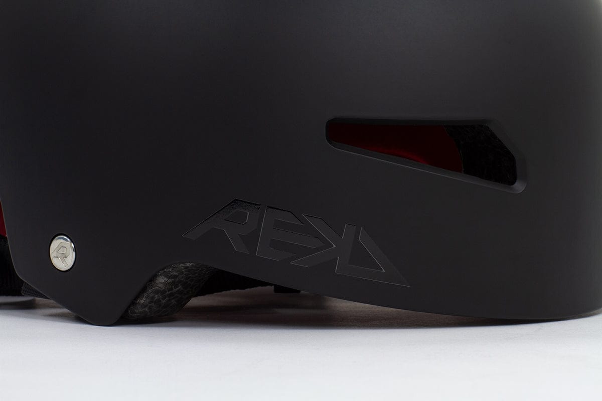 Close-up of the black REKD Elite 2.0 Skate/Scooter Helmet featuring the "REKD" logo and a ventilation slot. This certified action sports helmet highlights a metallic screw detail on the side, with its interior lining visible through the ventilation opening.