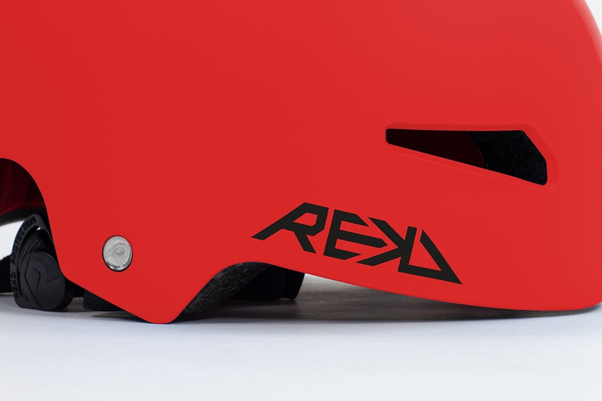 Close-up of a vibrant red REKD Elite 2.0 Skate/Scooter Helmet featuring a sleek black ventilation slot. The side prominently displays the "REKD" logo in bold black letters. Positioned on a light surface, this cutting-edge action sports helmet combines modern design with maximum safety and style.