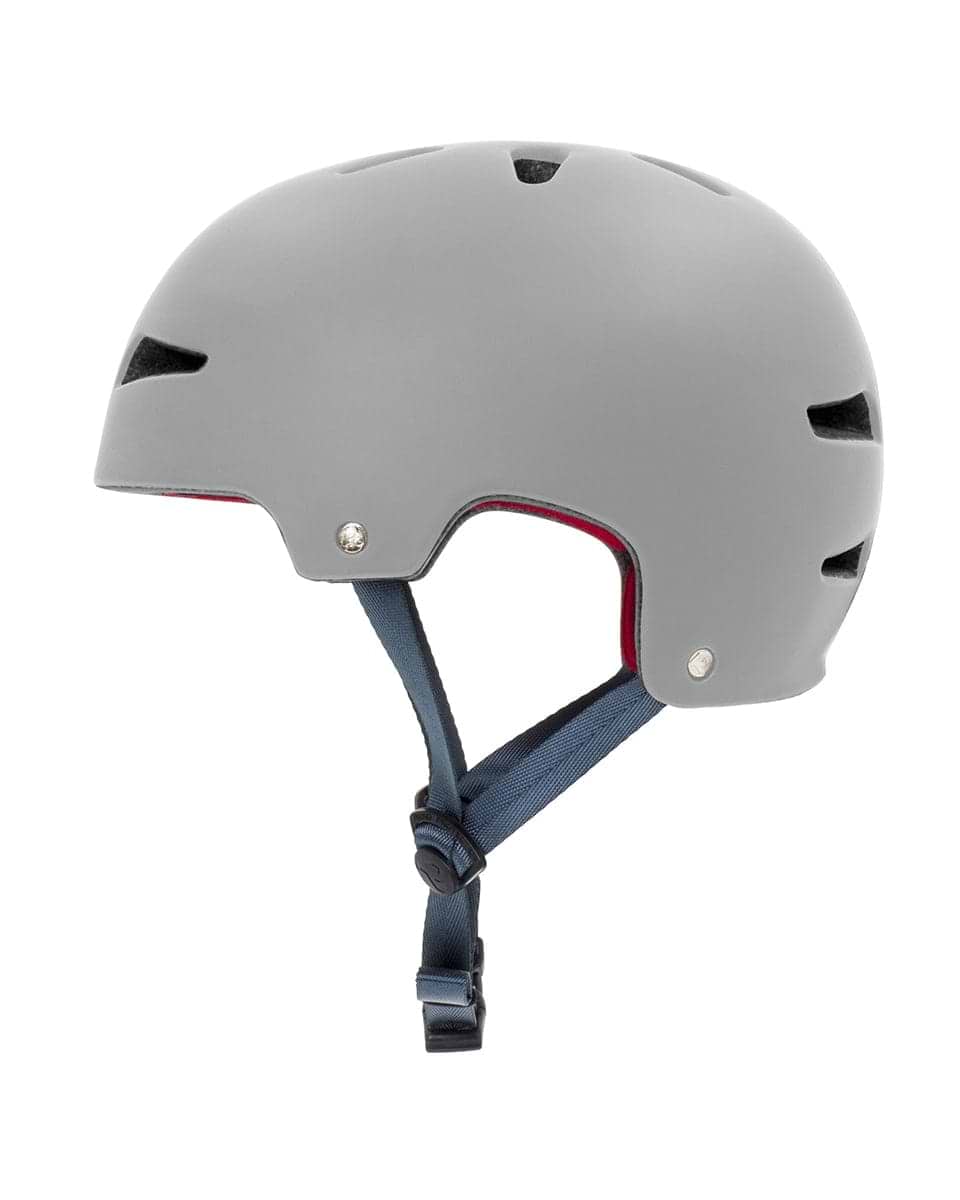 A side view of the REKD Ultralite In-Mold Skate/Scooter Helmet in grey highlights its matte finish. This helmet, designed for action sports, includes ventilation slots on the top and sides. It also features a blue adjustable chin strap with a black buckle. The advanced In-Mold technology is visible through the red interior lining, offering improved safety.