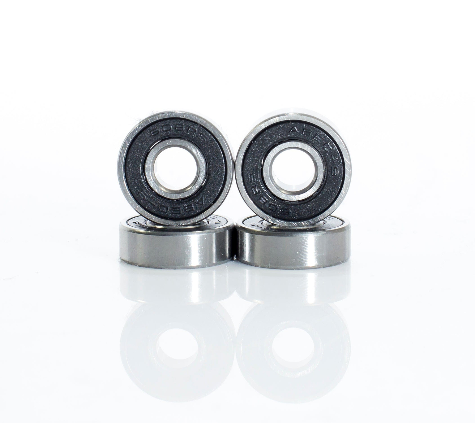 Four Skates.co.uk ABEC 9 Black Scooter Bearings, similar to those in a stunt scooter, are displayed in a staggered line with two standing and two lying flat. The metallic bearings showcase black inner rings against a simple white background, suggesting precision akin to their ABEC 9 ratings.