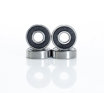 Four Skates.co.uk ABEC 9 Black Scooter Bearings, similar to those in a stunt scooter, are displayed in a staggered line with two standing and two lying flat. The metallic bearings showcase black inner rings against a simple white background, suggesting precision akin to their ABEC 9 ratings.
