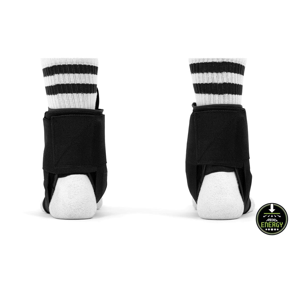 Rear view of two feet wearing REKD Energy Covert Ankle Skate Protection Braces in black over white socks with black stripes. A circular badge on the bottom right corner showcases the words "REKD Energy," highlighting enhanced ankle protection along with a stylized mountain graphic.