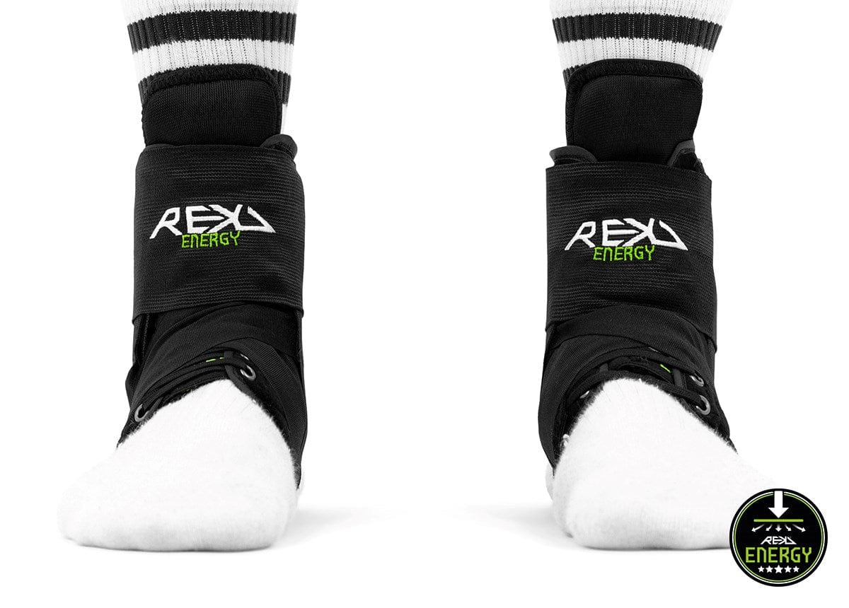 Close-up of a person wearing REKD Energy Covert Ankle Skate Protection Braces in black over white socks. The sturdy ankle braces provide coverage for the lower shins and feet. Above the braces, striped portions of the socks are visible, with a logo situated in the bottom right corner.