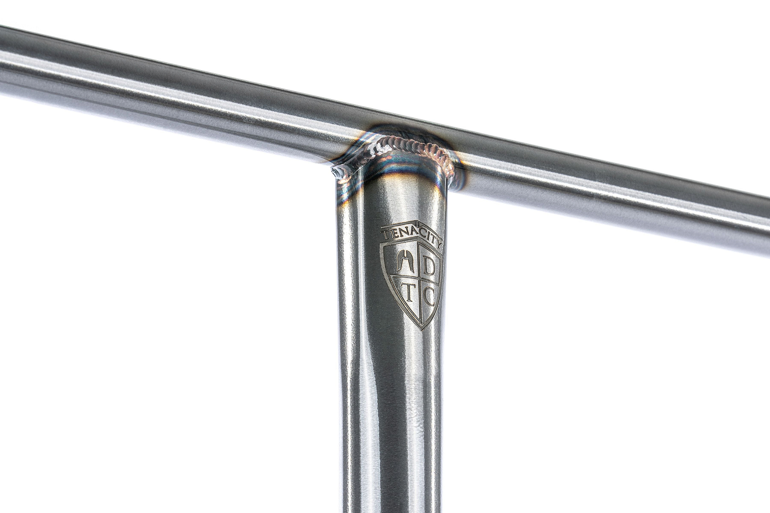 Close-up of a metal T-joint with visible weld marks and an engraved shield-shaped logo reading “TENACITY ABT.” The photo showcases the quality craftsmanship and metallic sheen of the Ethic DTC Tenacity V2 Steel Oversized HIC Stunt Scooter T-Bars (Raw, 770mm x 600mm) by Ethic, expertly constructed from durable chromoly steel to accommodate oversized scooter handlebars.
