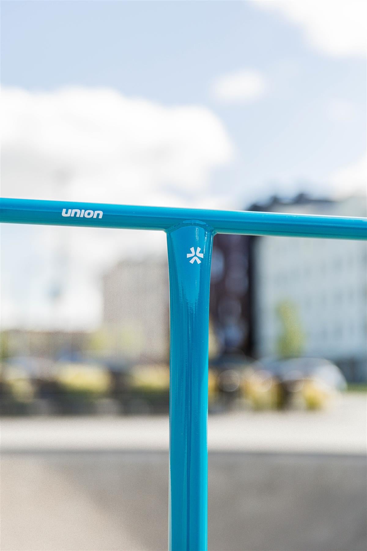 A close-up of the Union Uniq T Steel Standard IHC Stunt Scooter Bar in polished teal (550mm x 520mm), featuring the "Union" logo and a white symbol. The background is blurred with city buildings and a partly cloudy sky.