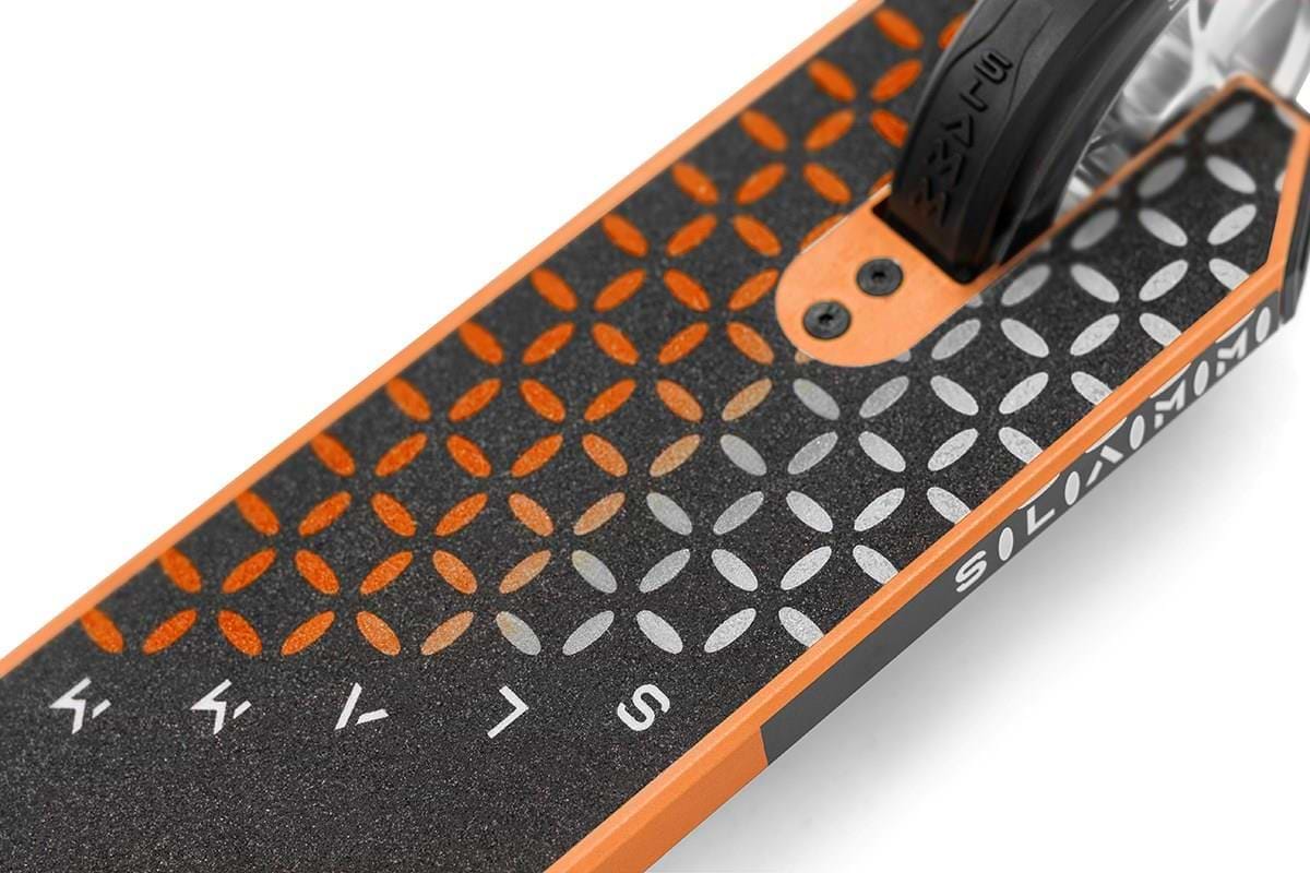 Close-up of the Slamm Assault Complete Stunt Scooter deck in orange, displaying a black and orange geometric pattern with white lettering, foot grip surface, and partially visible rear aluminum wheel—ideal for skill enhancement.