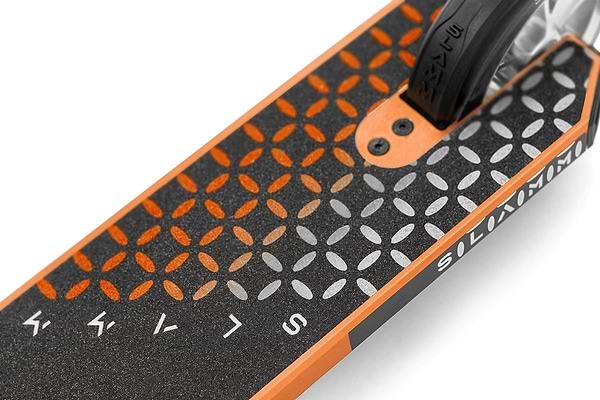 Close-up of the Slamm Assault Complete Stunt Scooter deck in orange, displaying a black and orange geometric pattern with white lettering, foot grip surface, and partially visible rear aluminum wheel—ideal for skill enhancement.