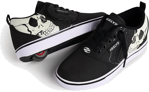 The Heelys Pro 20 PRNT Glow Boys Shoes in black/white feature glow-in-the-dark skull graphics, with one shoe displaying the low-profile wheel and the other showing off the bold skull design.