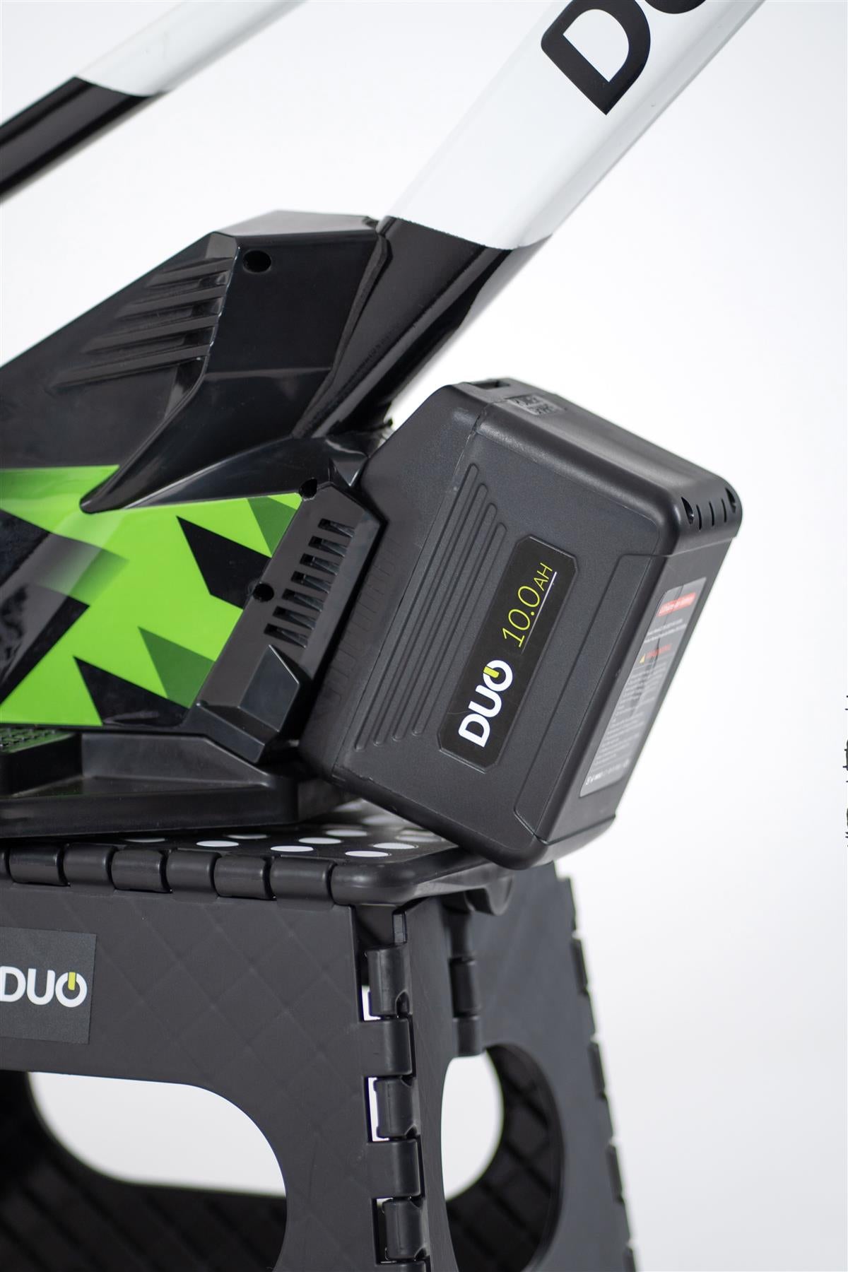 Close-up of a black 10.0Ah DUO battery on the Duo 20" Podium Pro Kids Electric Balance Bike in white and black, resting on a textured black plastic stand.