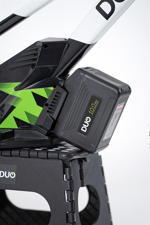 Close-up of a black 10.0Ah DUO battery on the Duo 20" Podium Pro Kids Electric Balance Bike in white and black, resting on a textured black plastic stand.