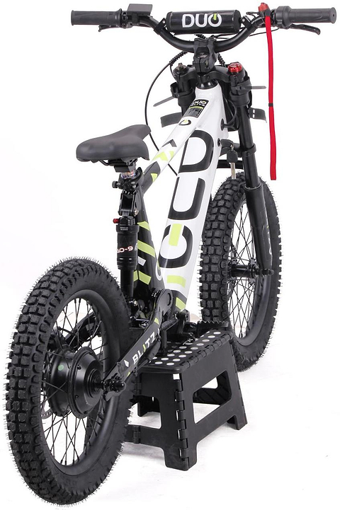 The Duo 18" Blitz Kids Electric Balance Bike in white and black, by Duo, sits on a black step stool. It features chunky tires, a padded seat, and a red strap on the handlebars, shown from the rear angle.