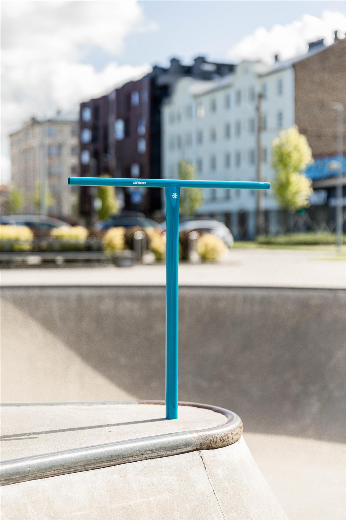A Union Uniq T Steel Standard IHC Stunt Scooter Bar in polished teal (550mm x 520mm) stands upright on a concrete skatepark edge, with blurred buildings, trees, and parked cars in the partly cloudy background.
