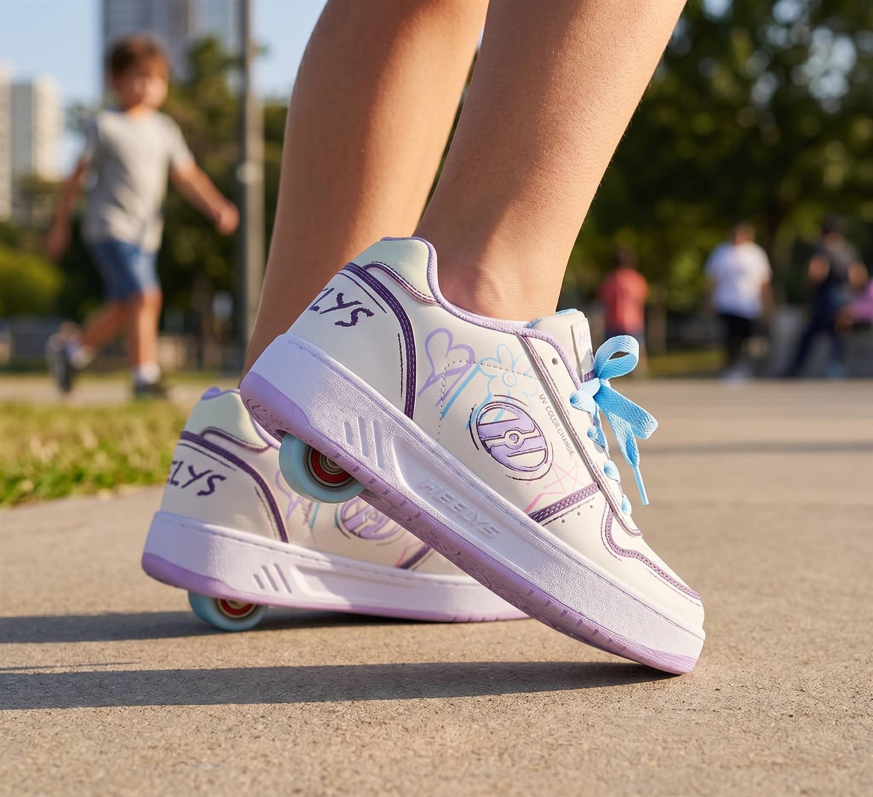 A person stands on a paved path wearing Heelys Kama Colour Changing Girls Shoes in white, lilac, and blue; wheels are visible in the soles. These standout Heelys roller shoes for kids are set against a blurred park background with people and trees.