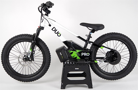 A Duo 20" Podium Pro Kids Electric Balance Bike in white and black with green accents stands on a small black stand against a plain white background. "DUO" and "PRO" are branded on the frame and battery.