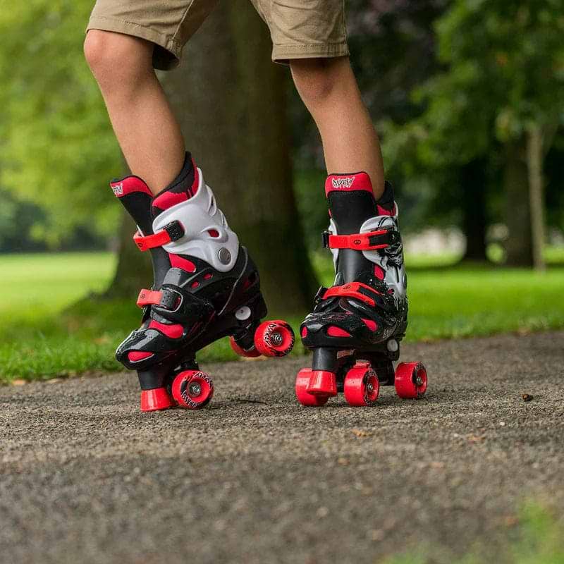 A child in tan shorts wears Osprey Skull Adjustable Quad Roller Skates - Red (by Osprey) on a paved park path, surrounded by grass and trees. These skates are ideal for kids learning to skate outdoors.