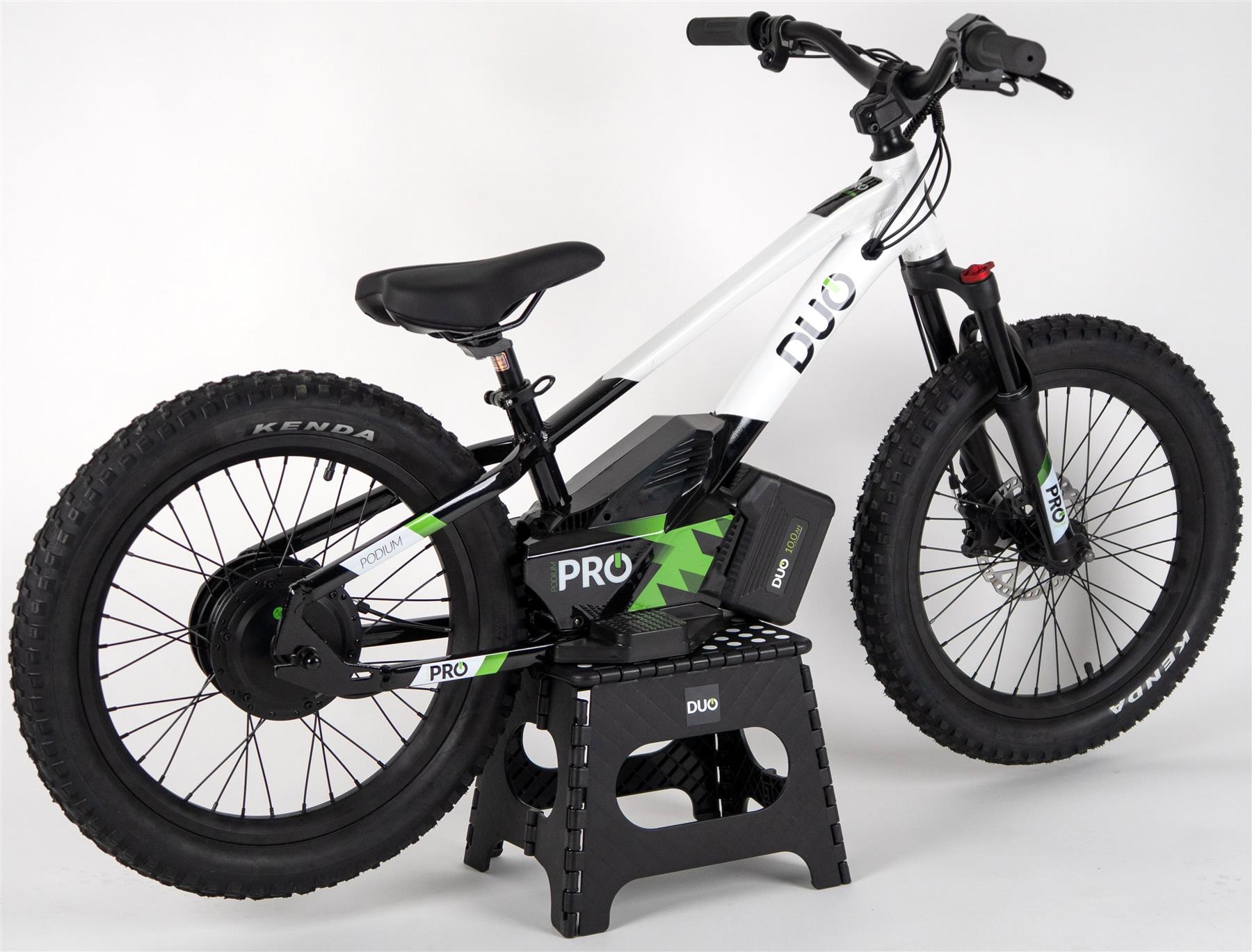 The Duo 20" Podium Pro Kids Electric Balance Bike in white and black, featuring thick tires and a battery pack for off-road use, is shown on a black stand against a plain white background.