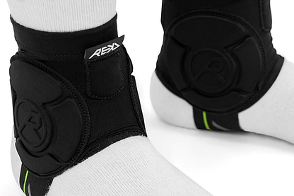 Close-up of two feet in white socks wearing REKD Energy Covert Ankle Impact Skate Protection Guards - Black, featuring Velcro straps, "REX" tag, padded energy-absorbing protection to reduce impact, and a green accent at the back.