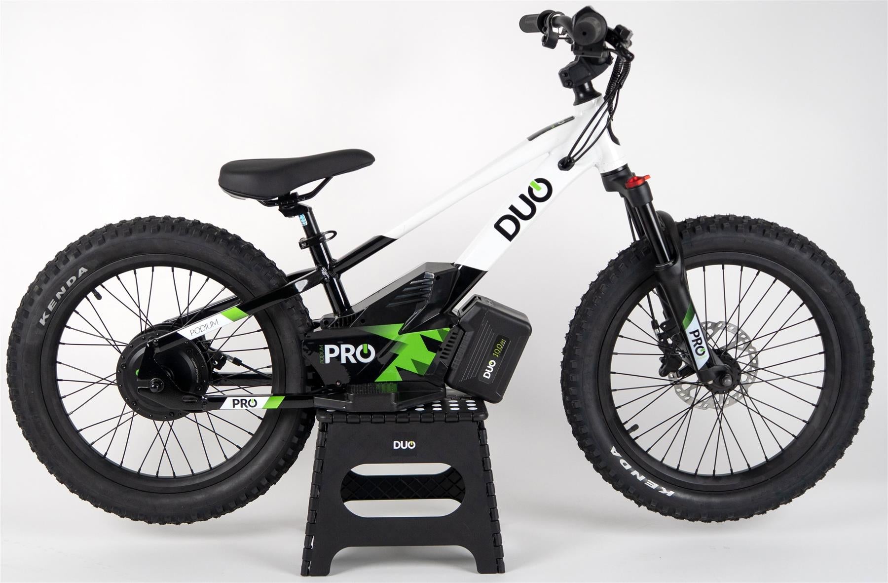The Duo 20" Podium Pro Kids Electric Balance Bike in white and black is shown on a black stand against a plain white background, featuring a visible battery, thick tires, and sturdy frame.