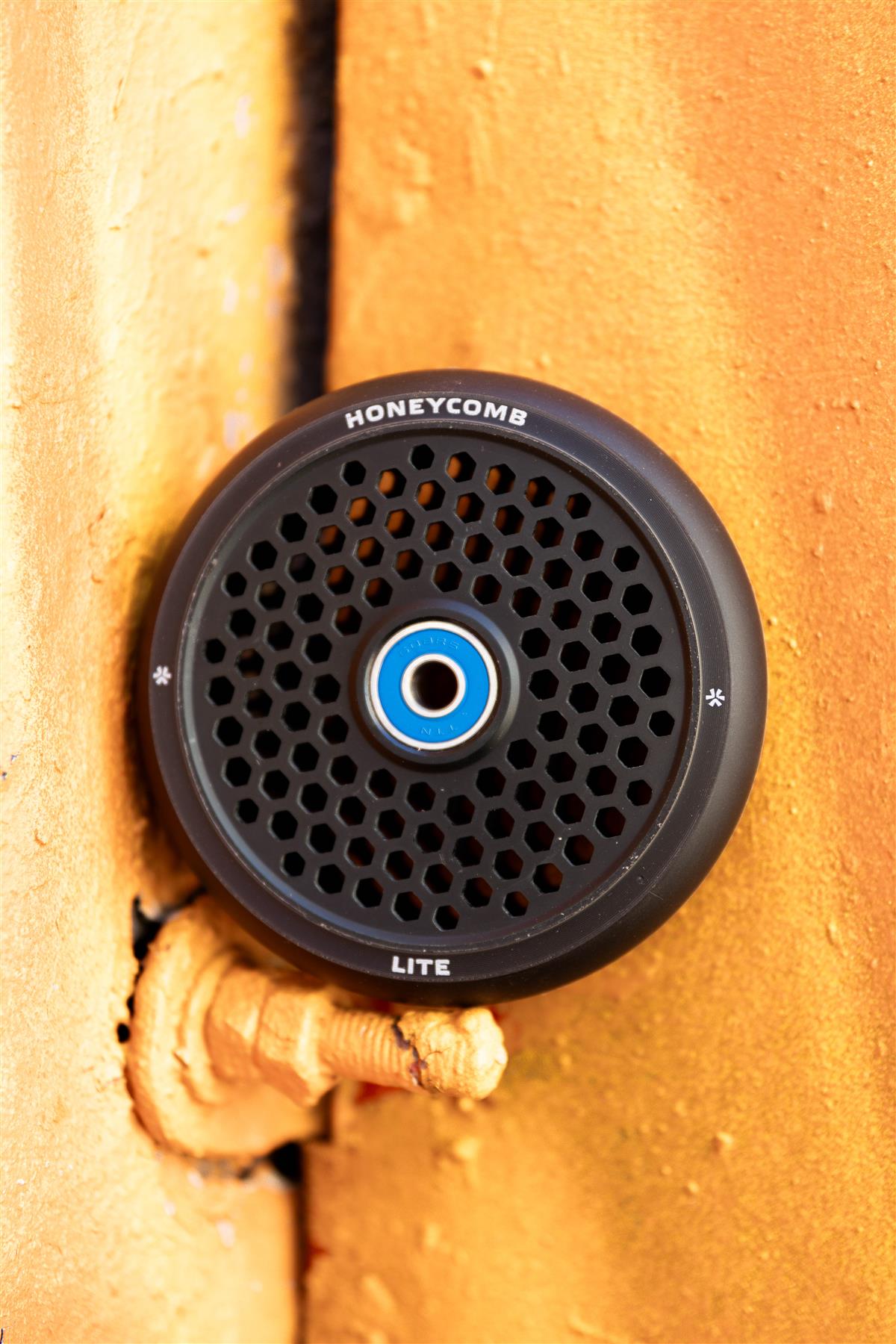 The Union Honeycomb Lite 110mm Stunt Scooter Wheel in black features a lightweight honeycomb grille design and is shown mounted on an orange textured surface with a visible pipe below.