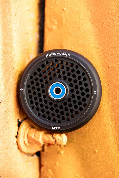 The Union Honeycomb Lite 110mm Stunt Scooter Wheel in black features a lightweight honeycomb grille design and is shown mounted on an orange textured surface with a visible pipe below.