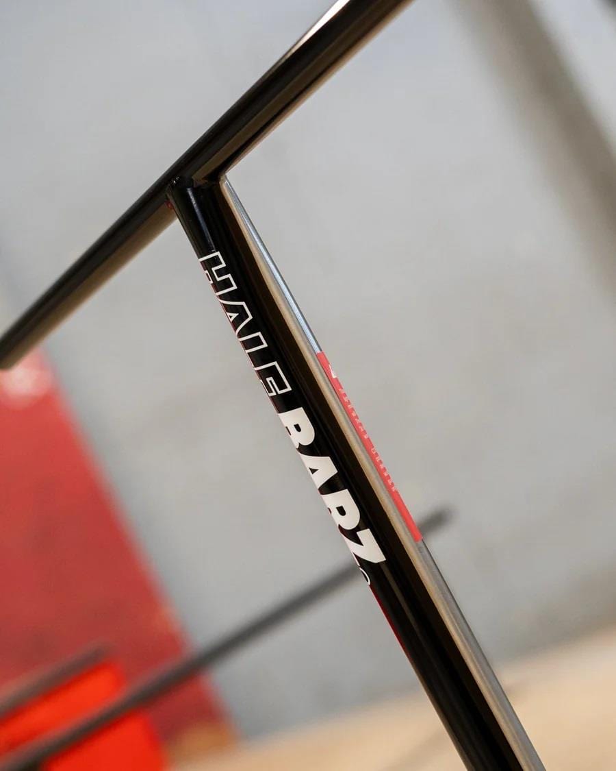 Close-up of a black and chrome T-bar handlebar with "CORE" etched in white, drawing inspiration from sleek stunt scooter designs. The blurred backdrop implies a lively and adventurous setting accented with red and gray tones, ideal for park riders looking for thrills.