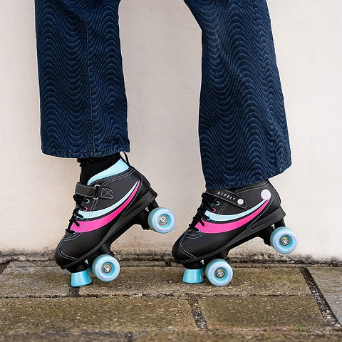 A person stands on stone pavement wearing Osprey Disco Quad Roller Skates - Black by Osprey, with only their lower legs and blue textured pants visible—ideal for indoor and outdoor skating fun.