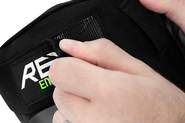 A person carefully adjusts the strap of black knee protection gear branded with the REKD logo. The hands secure a Velcro patch to ensure an optimal fit for extreme sports, enhancing impact absorption while wearing the REKD Energy Patrol Ramp Knee Skate Protection Pads in Black.