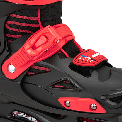 Close-up of Seba STJ Junior Adjustable Inline Skates in black and red, featuring a red buckle strap, black mesh with red accents, and a stars-and-stripes patch. Ideal for beginners; partial view of wheel and frame at the bottom.