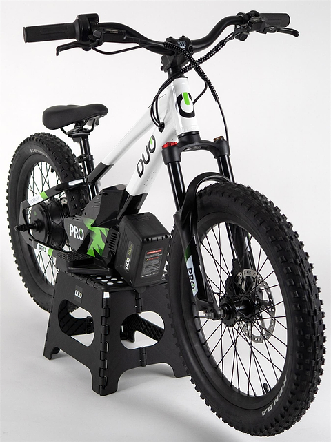 The Duo 20" Podium Pro Kids Electric Balance Bike by Duo, in white and black, features thick tires, green accents, and a visible battery pack. Displayed on a black stand with “DUO” on the frame, it’s perfect for kids seeking an electric balance bike.