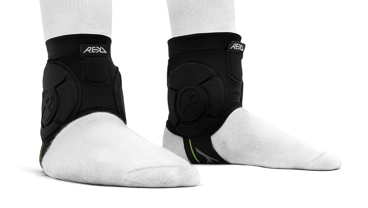 Two feet wear white socks and REKD Energy Covert Ankle Impact Skate Protection Guards in black, shown side and front. The REKD-branded supports feature straps and energy-absorbing protection for skating.