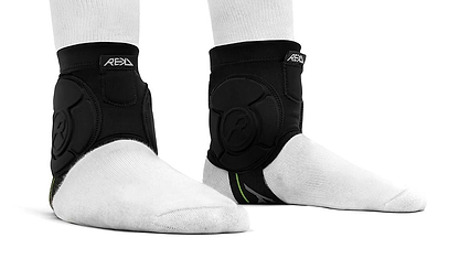Two feet wear white socks and REKD Energy Covert Ankle Impact Skate Protection Guards in black, shown side and front. The REKD-branded supports feature straps and energy-absorbing protection for skating.