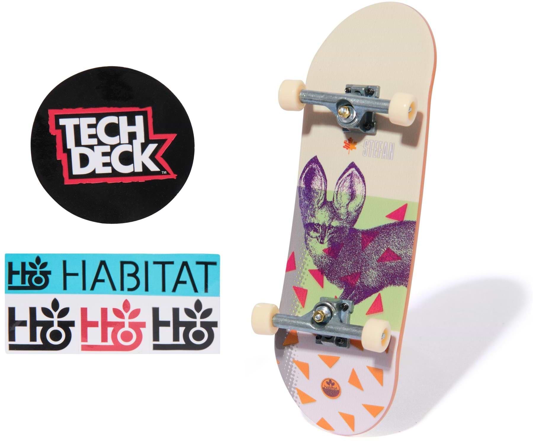 A Tech Deck 96mm M57 Fingerboard - Habitat stands upright, featuring a purple bat and orange triangles. Nearby are a round Tech Deck sticker and three rectangular Habitat logo stickers in blue, black, and red.