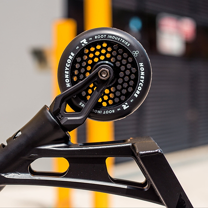 Close-up of the Root Industries Honeycore 110mm Stunt Scooter Wheel in Black, attached to a scooter frame with yellow bollards and a blurred building in the background.