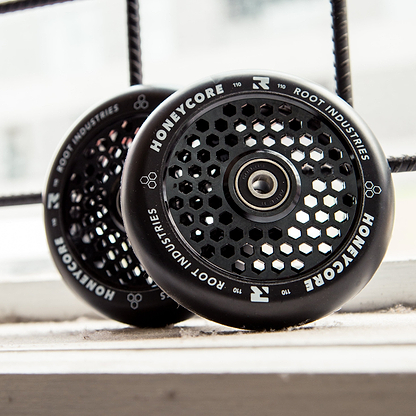 Two Root Industries Honeycore 110mm Stunt Scooter Wheels in black, featuring hexagonal cutouts and distinctive branding, rest on a white windowsill with a metal grid and cityscape blurred in the background.