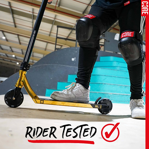 Person wearing black knee pads and white sneakers stands on a gold scooter with CORE Hollow Core V2 100mm Stunt Scooter Wheel - Black at a skate park. Blue stairs, curved ramp, and “RIDER TESTED” text with red checkmark behind them.