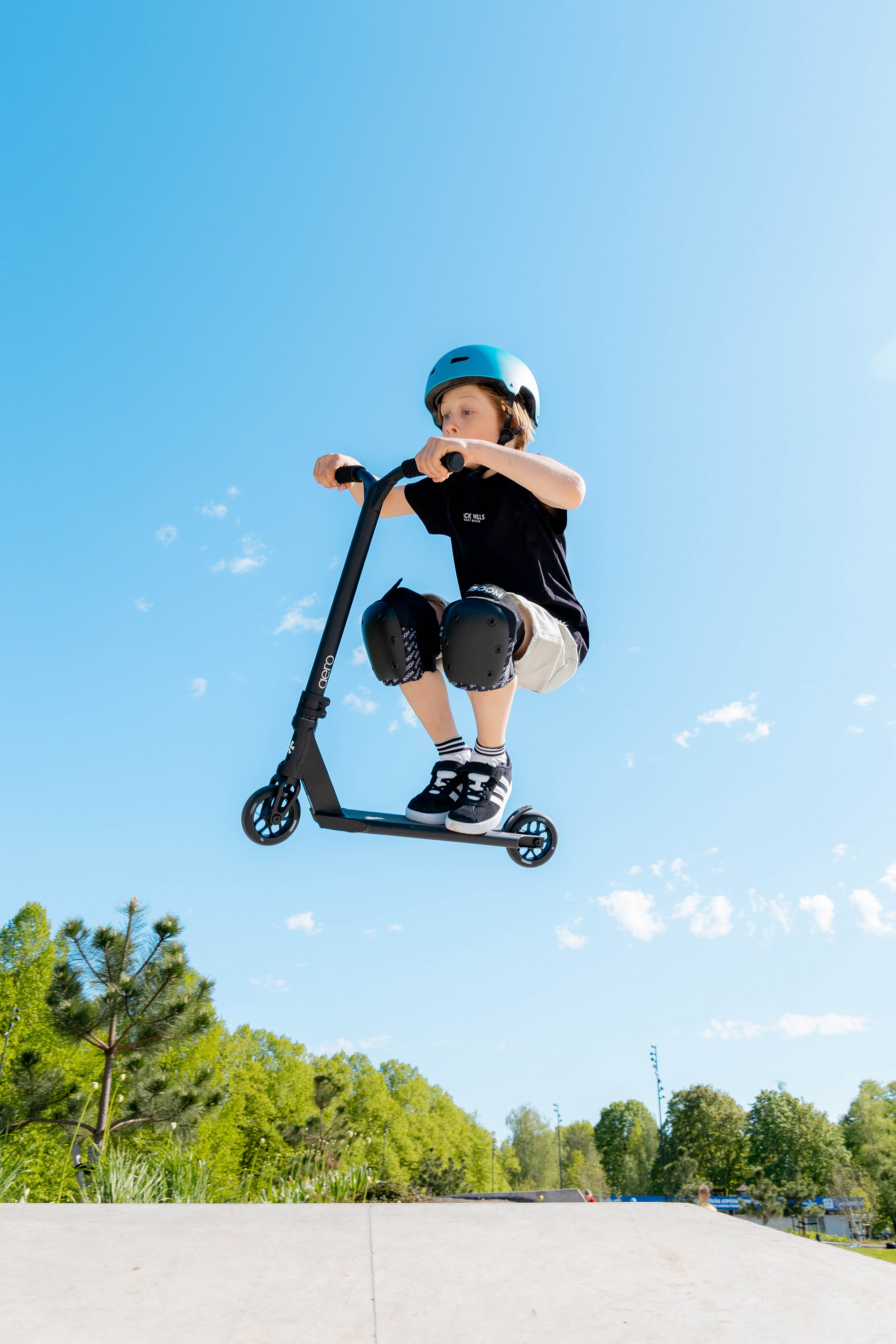 A young boy in a blue helmet and knee pads performs a jump on the Union Aero Complete Stunt Scooter - Black by Union at a skatepark, with trees and a clear blue sky in the background.