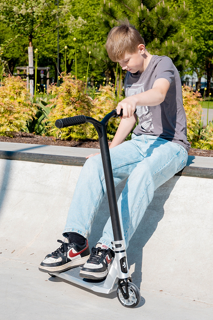A boy in a gray t-shirt and light blue jeans sits on a concrete ledge at a skatepark, holding the Union Aero Complete Stunt Scooter - Black / Raw by Union. Green plants and trees are visible behind him.