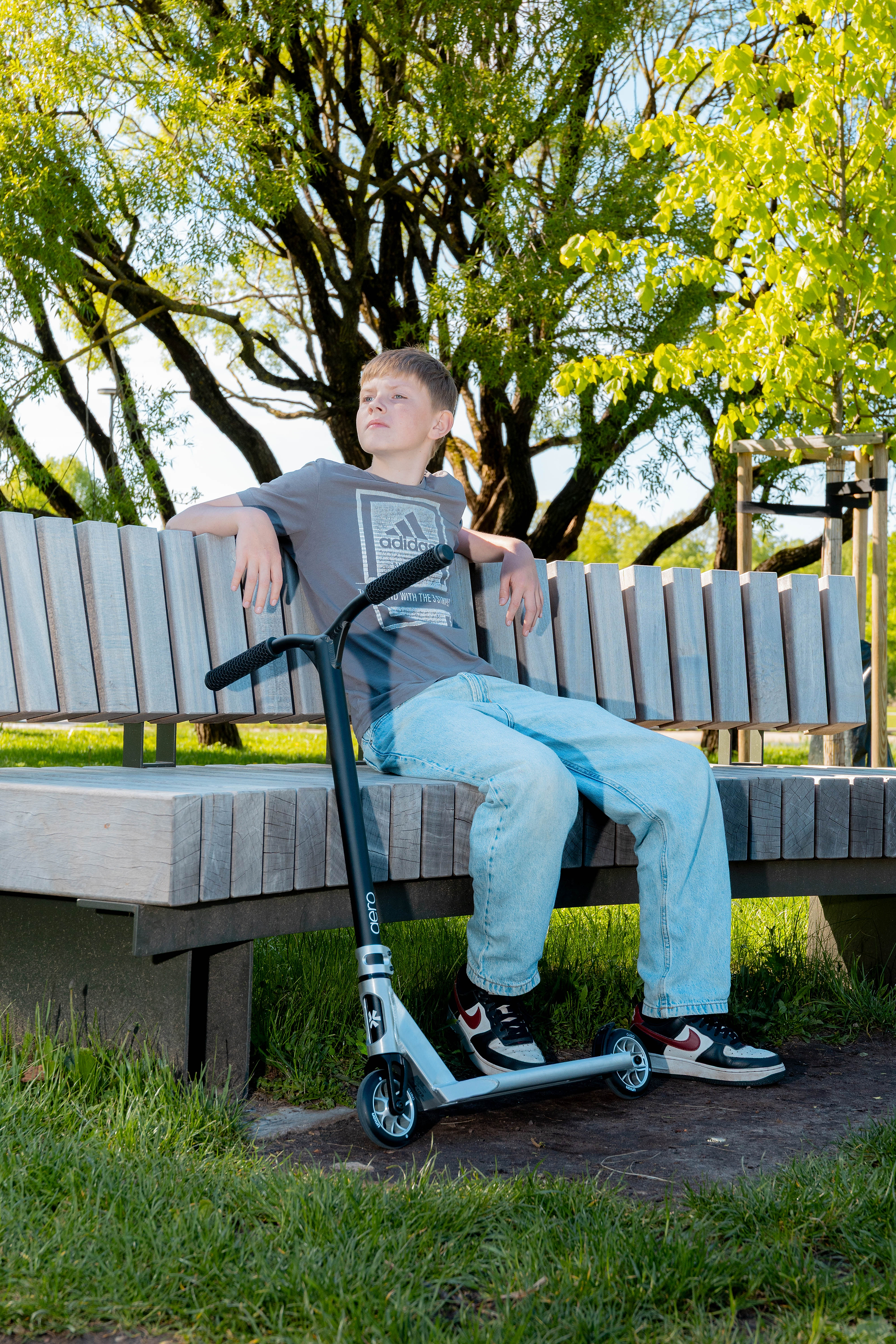 A boy sits on a park bench holding the handlebars of a Union Aero Complete Stunt Scooter by Union in Black / Raw, looking to the side with green trees and sunlight in the background.