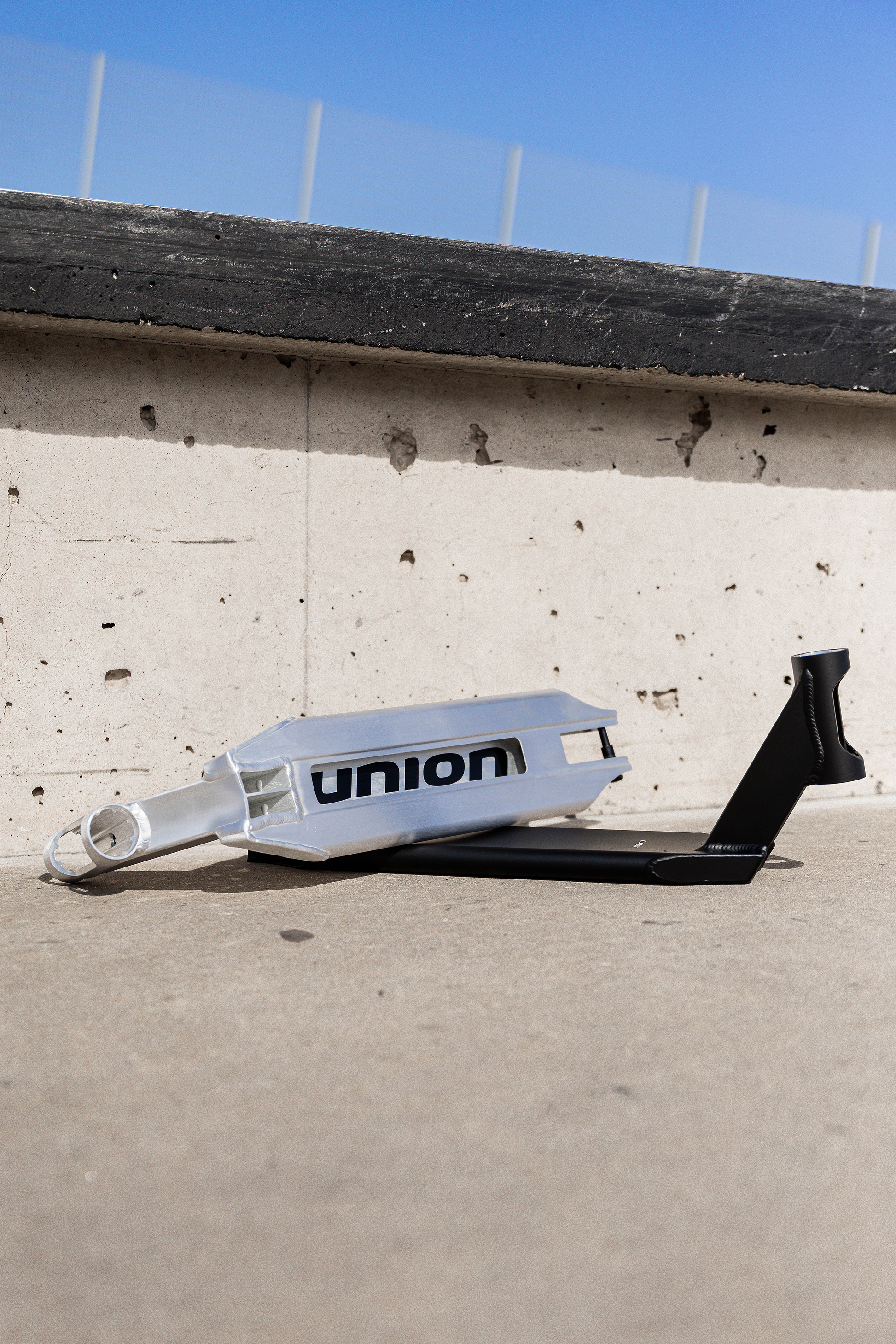 The Union Classic Pro Silver Stunt Scooter Deck - 4.75" by Union, with "UNION" on the side, sits on concrete and leans against a light wall under a blue sky.