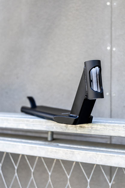 The Union Classic Pro Black Stunt Scooter Deck - 4.75" from Union rests on a metal railing in front of a gray textured wall, showcasing its smooth edges and angular design.
