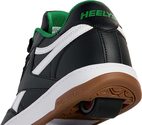A close-up of the Heelys x Reebok Classic Boys Shoes - Black / White / Gum shows a black shoe with white sole, gum accents, green inner lining, visible heel wheel, and Heelys branding on the heel and sole.