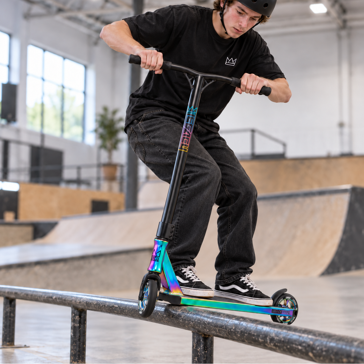 A rider in a helmet and black attire balances the Legacy Hype Complete Stunt Scooter - Rainbow Neochrome by Legacy on a rail at an indoor skatepark, with ramps and platforms visible in the background.
