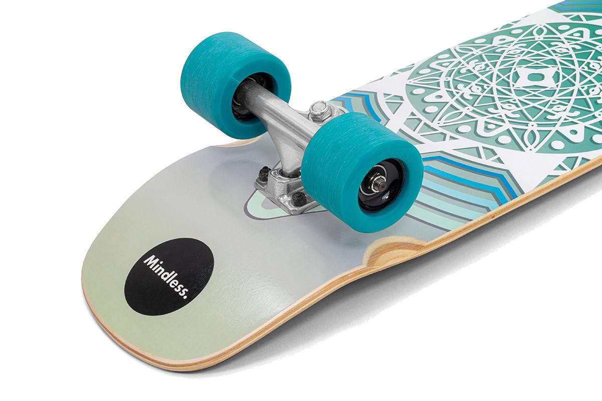 A close-up of the Mindless Drifter Cruiser Skateboard 28″ x 8″ - Teal shows teal wheels, silver trucks, and geometric teal and white designs, with “Mindless” on a black circle at the tail.