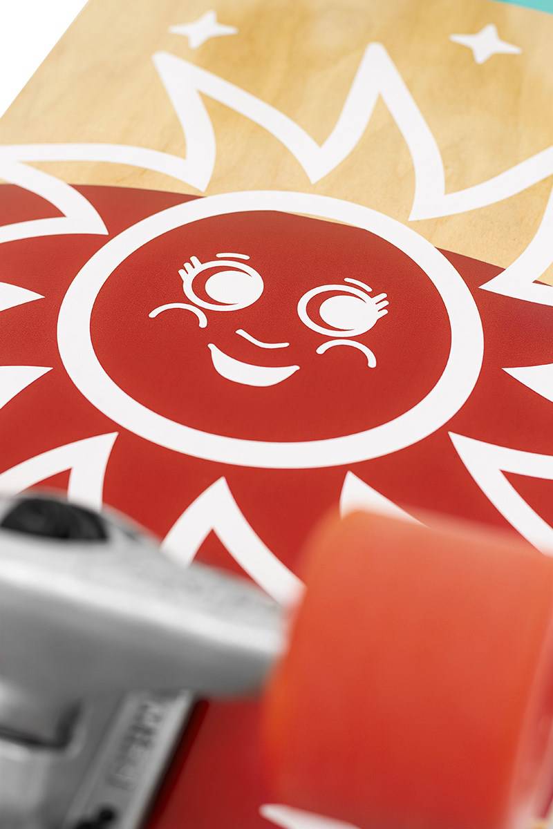Close-up of the Mindless Soul Surf Skate Cruiser Skateboard 29.5″ x 9.75″ - Red, showing its red sun graphic with a smiling cartoon face, orange wheels, and metal truck in the foreground. Brand: Mindless.