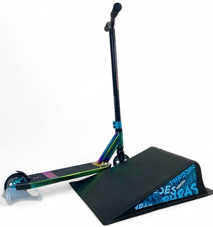 A colorful stunt scooter balances its front wheel on the Phrases One Way Kicker Skate Ramp by Phrases, featuring blue text on the side, set against a plain white background.